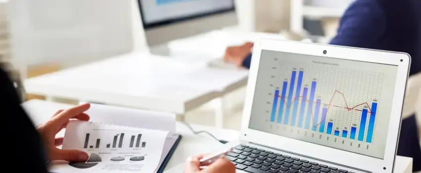 Un profesional de marketing analiza en detalle informes y gráficos de métricas publicitarias digitales en una computadora portátil y documentos físicos, destacando la importancia de la toma de decisiones basada en datos.
