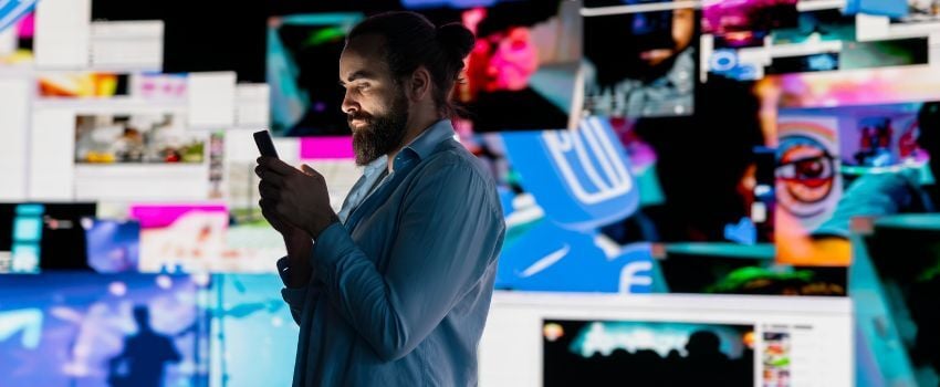 Persona navegando en su celular con diversos medios y canales reflejándose en una pantalla detrás, ejemplo de la transición de un usuario entre narrativas transmedia