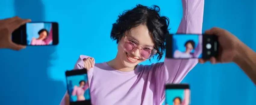 Joven creadora de contenido sonriendo y posando con gafas de sol frente a múltiples teléfonos móviles que la graban, ilustrando el éxito y la viralidad en redes sociales sobre un fondo azul.